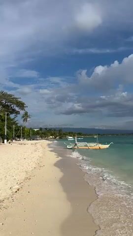 A vertical shot of a beach on a bright, cloudy day. The sand is light beige and curves gently into the turquoise water. The water is clear and shallow near the shore, with small waves lapping at the sand. A yellow and white boat with a thatched roof is anchored just offshore. In the background, palm trees and other greenery line the coast, with buildings and hills visible in the distance. The sky is a vibrant blue with scattered white and grey clouds.