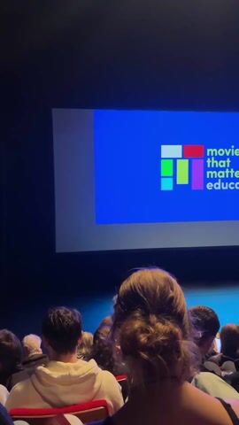 The video shows a room full of people sitting in red seats facing a large screen displaying a blue background with the words "movies that matter educate" and a colorful block-like design. The camera is positioned behind several audience members, partially obscuring the view of the screen at times. The room appears to be a small theater, dimly lit. The audience consists of young adults. Some are talking quietly, while others seem to be focused on the screen. The video is a bit shaky and the focus shifts throughout.