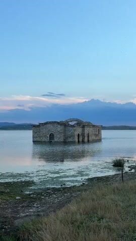 This media file captures the striking image of the submerged St. George's Church in Panicherevo, Bulgaria. The stone structure stands partially submerged in the calm waters of Lake Zhrebchevo, its weathered walls and arched windows a testament to its history. The church, appearing as an isolated island, is the sole subject of the photograph.

The scene is set during dusk, with the sky painted in hues of soft blue and delicate pink, transitioning from the daylight to twilight. Wispy clouds drift across the horizon, adding depth to the serene atmosphere. The water's surface reflects the muted colors of the sky and the silhouette of the church, creating a tranquil and somewhat melancholic ambiance. The surrounding landscape is subtly visible in the distance, with rolling hills under the vast expanse of the sky.

The foreground features the shoreline, covered in dry grass and pebbles, suggesting a natural, untouched environment. The water itself shows patches of greenish aquatic vegetation, adding a touch of life to the submerged scene. The overall mood is one of peaceful solitude and quiet contemplation, evoking a sense of lost time and submerged memories. No people are visible in the frame, emphasizing the solitary nature of the church. There is no visible text in the media file.