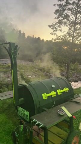 A barrel smoker sits outdoors in a grassy area, with smoke billowing from its chimney. The smoker is dark green with bright yellow accents on its doors and a Texas license plate reading "GPG-2985". A metal cart with wheels is attached to the smoker, holding a small container and some papers. A bucket hangs from the smoker. In the background, a rocky riverbed leads to a wooded area with trees. The sky is a warm orange and yellow, suggesting either sunrise or sunset. The overall atmosphere is peaceful and serene, possibly a quiet evening at a campsite or backyard.