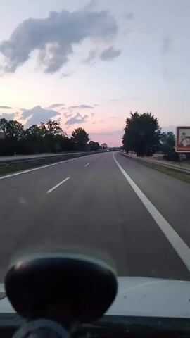 This media file shows a point-of-view shot from inside a vehicle driving on a highway. The highway has multiple lanes marked with white lines. Trees and vegetation line the sides of the road. In the distance, other vehicles can be seen with their headlights on. The sky is a dusky pink and purple, with clouds scattered across it, suggesting it is either dawn or dusk. The overall mood is peaceful and serene, with the fading light casting a soft glow over the landscape. The location is identified as Illertissen, Germany. There is no visible text in the media file.