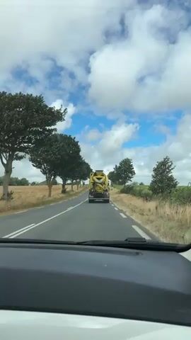 The video shows a rear view of a yellow concrete mixer truck driving down a two-lane paved road. The truck has a large cylindrical drum on its back and a chute extending from the drum. The truck has a license plate that reads CT 533 SJ. On the back of the truck, there are yellow speed limit signs indicating 80 and 90. The road is lined with trees on both sides and fields beyond the trees. The sky is cloudy with patches of blue. The video appears to have been filmed from inside another vehicle, as the dashboard is visible in the foreground.