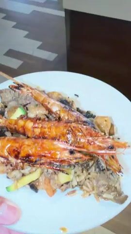 A close-up shot shows a white plate filled with food, including rice, meat, vegetables, and three large grilled shrimp on skewers. The camera then pans to the right, revealing a buffet area with various food pans. A person is seen from the back, reaching towards the buffet. The camera then pans further to the right, showing a young boy selecting food from the buffet. The environment appears to be a restaurant or dining hall with wooden floors and a buffet setup.