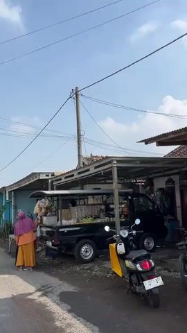 The video shows an outdoor market scene. Two individuals are visible, one wearing a purple headscarf and an orange top with a yellow skirt, the other is partially obscured by a black pickup truck. The pickup truck is loaded with various goods, including what appear to be vegetables and other food items. A canopy or awning extends over the truck, providing shade. A utility pole with wires is prominent in the background, against a clear blue sky. The ground appears to be a dirt or gravel road.