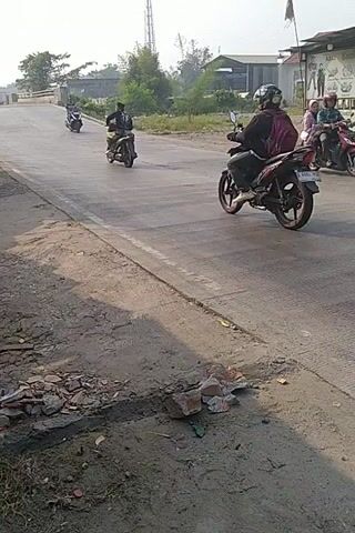 The video shows a winding paved road with motorcycles traveling on it. The road is bordered by a dirt embankment on the right and vegetation and buildings on the left. The lighting suggests it is daytime. Several motorcycles with riders are visible at different points along the road. One motorcycle appears to be a scooter, while others are standard motorcycles. The video is shot from a low angle, possibly from another motorcycle or a vehicle. The overall scene is one of casual motorcycle activity on a rural or semi-rural road.