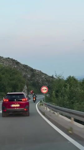 The video shows a red Peugeot SUV driving on a paved road that curves gently to the left. The car is followed by a motorcycle with a red flag attached to it. The road is bordered by a metal guardrail on the right and green vegetation on the left. A road sign indicating "no overtaking" is visible next to the guardrail. In the background, there is a mountainous landscape under a dusky sky. The SUV's brake lights illuminate at one point.