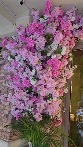 The video shows a decorative arrangement of artificial pink and white flowers, resembling cherry blossoms, adorning an indoor space. The flowers cascade down a wall and over an archway, creating a vibrant and whimsical atmosphere. The scene is illuminated by soft indoor lighting, and the time of day is unclear.

The background features a brick wall on the left and a glass door on the right. Through the glass door, a reflection of an outdoor scene is visible, with cars and buildings suggesting an urban or suburban setting. On the door itself, there are some pictograms and text that appear to be in Cyrillic. One visible sign reads "ШАНОВНІ ГОСТІ," which translates to "Dear Guests" in Ukrainian.

A security camera is mounted on the ceiling in the upper left corner of the frame. In the foreground, a large potted plant with green, spiky leaves is partially visible. The overall impression is that of a stylishly decorated interior, possibly a cafe, restaurant, or shop, designed to create a welcoming and visually appealing ambiance for visitors. The abundance of flowers evokes a sense of springtime and natural beauty, even though they are artificial. There are no people visible in the video.