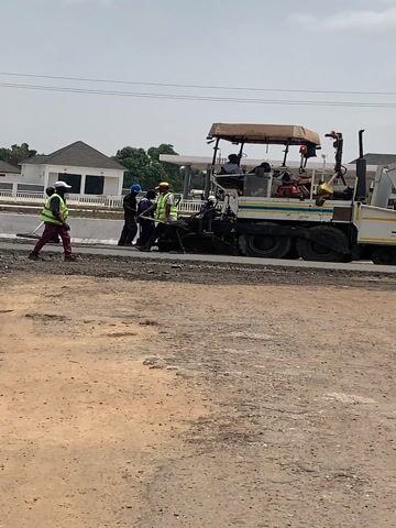 The video captures a dynamic scene of road construction taking place on a bright, sunny day. In the mid-ground, a large asphalt paver, predominantly white and yellow with a beige canopy over the operator's area, is slowly moving from left to right across the frame. An operator is visible in the driver's seat of the paver.

Several construction workers are actively involved in the process, wearing high-visibility vests (yellow and green) and some with hard hats, indicating safety measures are in place. On the left side of the paver, a worker in a yellow vest and maroon pants is seen walking forward, holding a rake or a similar tool, seemingly leveling fresh asphalt. Other workers are positioned around the paver, assisting with the laying and spreading of the asphalt.

The ground in the immediate foreground is unpaved, consisting of rough, dusty dirt and small stones, contrasting with the smooth, dark asphalt being laid down by the machine. In the background, modern-looking residential buildings with light-colored walls and dark roofs are visible under a clear, slightly hazy sky, suggesting a suburban or recently developed area.

At approximately the 0:02 mark, a dark-skinned hand briefly enters the frame from the bottom, fingers spread wide open, partially obscuring the view of the construction activity. The hand then closes into a fist and exits the frame around 0:04, bringing the construction scene back into full focus.

As the video progresses, the asphalt paver continues its work, with the workers moving in coordination around it. The side of the paver clearly displays the letters "JRB" followed by what appears to be a smaller "a" and another partially visible word starting with "c", possibly "construction". Below this, another word, "LAGOS", might be present but is difficult to discern due to blurriness. The continuous sound of heavy machinery accompanies the visual of the ongoing road work.