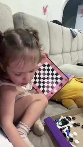 A young girl with brown hair in pigtails sits on a gray couch, wearing a white tank top and pink socks. She is holding a pink and black checkered board. In front of her is a box filled with chess pieces, some black and white, and a purple case. To her right, a yellow stuffed animal rests on the couch. The background shows a cream-colored wall with some red markings. The girl looks towards the camera and then waves her hand in front of her face.