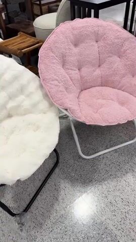 The video shows a close-up of two fluffy, round chairs, one white and one pink, in what appears to be a retail store. The chairs have metal frames and plush, soft fabric. The white chair has a black frame and the pink chair has a white frame. In the background, other furniture and merchandise are visible, including shelves with clothing and decorative items. The camera pans slightly, showing more of the store's interior and hinting at a wider selection of chairs, including black and white options in later frames. The lighting suggests an indoor environment, possibly a department store or home goods store. There are no people clearly visible interacting with the chairs.