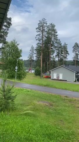 The video shows a residential property with a light green, single-story building in the background. Two white doors are visible on the building, along with two white pipes extending from the roof. Several red kayaks are leaning against the side of the building. A paved pathway runs in front of the building and curves to the left. Tall pine trees are scattered throughout the scene, some reaching above the building. In the foreground, lush green grass and a small evergreen tree are visible. A signpost with a white circular sign is located to the left of the pathway. In the distance, a body of water is visible, with what appear to be recreational structures, possibly a playground or park area, on the opposite shore. The sky is overcast with clouds.