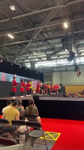 A group of performers, mostly children, dressed in traditional Japanese attire, stand on a stage. Some are wearing red robes and black pants, others are in colorful shirts. They appear to be in the middle of a performance, possibly a dance or a musical act, as some are holding instruments like drums. A large audience is seated in chairs in the foreground, facing the stage. The environment is an indoor venue with a high ceiling and industrial lighting. The stage is decorated with banners and has a backdrop with colorful graphics. The overall scene suggests a cultural event or festival.