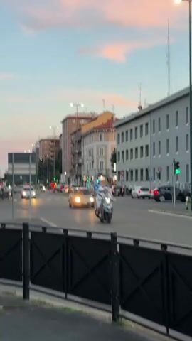 The video captures a street scene at dusk. Cars and a scooter are driving on a road lined with buildings. The sky displays soft pink and blue hues from the setting sun, with a few wispy clouds. Traffic lights are visible, with some showing green. Pedestrians are crossing a street at a marked crosswalk, and a person is riding a bicycle. A black metal fence is in the foreground, obscuring the bottom portion of the view. The overall atmosphere is tranquil and urban.
