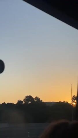The video shows a view from inside a vehicle of a sunset over a landscape. The sky is a gradient of blue to orange. Trees are visible in the foreground and midground. A white van is driving on a road in the lower left corner. A street light is visible in the center of the frame. The sun is low on the horizon, casting long rays of light. The view is framed by the interior of the vehicle, with a dark curtain on the right and a dark window frame. A person's head is partially visible in the lower right corner of the frame.