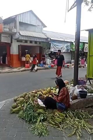 The video shows a street scene in front of a market. A person is sitting on the sidewalk next to a pile of fruits and vegetables. A man in a black shirt and a red sarong walks past. Another person on a motorbike is seen in the background. The market stalls are visible behind the people. The scene appears to be a typical street market environment.