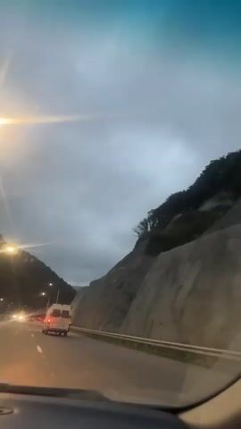 The video shows a view from inside a vehicle traveling on a highway at dusk or in overcast conditions. The road is flanked by a steep, rocky hillside on the right and a more densely vegetated hillside on the left. Several vehicles are visible on the highway ahead, with their headlights and taillights illuminated. The most prominent vehicle is a white van driving in the same direction as the viewpoint vehicle. Streetlights are also visible along the road, casting a yellow glow. The sky is cloudy and appears to be either early morning or late evening. The overall impression is of a journey on a highway in a mountainous or hilly region under dim lighting conditions.