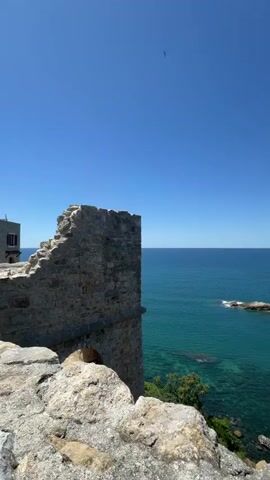 The video opens with a view of the sea and a stone wall, then pans to reveal a coastal town with buildings nestled into a hillside. Rocks are visible in the turquoise water. The camera then moves to a woman standing by a stone wall, looking at her phone. She has a red backpack and is wearing a yellow top and denim shorts. Behind her, more of the stone structure and a road with parked motorcycles are visible. The video ends with a shot of several parked scooters and motorcycles in front of the stone structure.