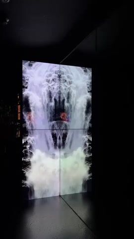 A video displays a large, vertical, four-panel screen showing a digital waterfall animation. The animation is symmetrical, with a central divide. The waterfall appears to be flowing downwards, with white and purple water and mist. In the center of the screen, on either side of the vertical divide, there are two bright orange objects that resemble abstract shapes or figures. The screen is placed in a dark room, with reflections visible on the floor. The lighting is dim, with the screen being the primary light source.
