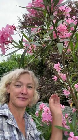 A woman with short blonde hair, wearing a plaid shirt, smiles at the camera. She is standing outdoors, in front of a tree with pink flowers and green leaves. The background shows more trees and greenery. The woman's right hand is visible, touching the tree.