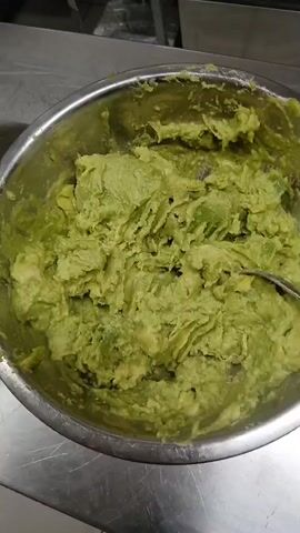 The camera briefly pans over two metal bowls sitting on a stainless steel surface, providing a top-down view. The bowl on the left contains a chopped mixture of green herbs, diced red tomatoes, and white onions. The bowl on the right contains mashed green avocado.