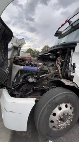 The video pans over the engine of a white semi-truck with the hood open. The engine is visible in detail, showing various components like hoses, wires, and metal parts. The truck is parked outdoors with a cloudy sky overhead. The front tire of the truck is also visible in the foreground. The video appears to be a close-up inspection or showcase of the truck's engine.