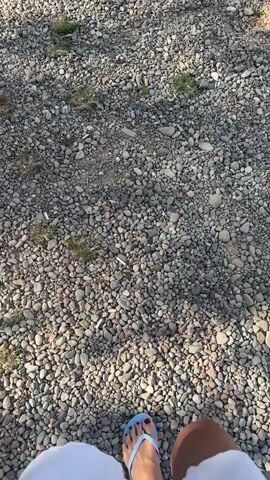 The video shows a first-person perspective of someone walking on a path covered with small, multi-colored pebbles. The person is wearing blue flip-flops with black nail polish on their toes. A portion of a white garment, likely a dress or skirt, is visible in the lower part of the frame, trailing behind them. Patches of green grass are scattered amongst the pebbles. The lighting suggests it is daytime, possibly late afternoon due to the long shadows. The camera is angled downwards, focusing on the person's feet and the ground.