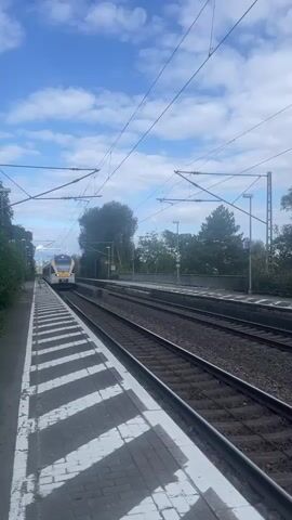 The video shows a train arriving at a station. The train is silver and yellow and has the word "eurobahn" written on its side. It is traveling on a set of train tracks. The station platform is visible in the foreground, with black and white striped markings. Overhead power lines for the train are also visible, along with trees and a blue sky with some clouds. The camera is positioned on the platform, looking down the tracks. The train moves from the distance towards the camera, passing by the platform and then continuing out of frame.