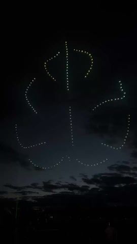 A nighttime shot shows a lighted figure of a shamrock in the sky, apparently formed by drones. There are also clouds in the sky. In the foreground, the silhouettes of onlookers are visible.