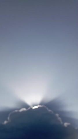 The video shows a time-lapse of a sun obscured by a cloud. The sun rays are visible, fanning out from behind the cloud, which is dark and textured. The sky is a clear, pale blue, gradually transitioning to a slightly darker shade towards the top of the frame. There are no people or objects other than the cloud and the sun visible in the video. The overall scene is one of atmospheric phenomena and the interplay of light and shadow in the sky.