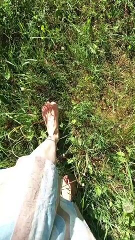 A first-person point of view captures a person's bare feet walking across a grassy field. The person is wearing a light-colored, striped skirt that reaches down to their ankles. The grass is green with some patches of brown. The sun is shining, and the light is bright.