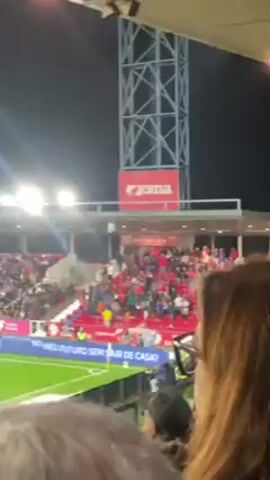 The video shows a crowd of people at a sports stadium, likely for a soccer game. The crowd is in the stands, wearing red. The stadium has a metal structure on the side, with a Joma sign on it. The sky is dark and lit up by stadium lights. People are cheering and excited. The video appears to be taken from within the crowd. The roof of the stadium is visible above the crowd.