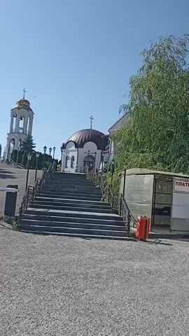The video opens on a clear day with a bright blue sky, showing a wide shot of a paved area leading up to a set of stairs. On the left side of the stairs, a white church steeple with a golden dome is visible. The stairs are made of stone and have metal railings on either side, leading up to a small white building with a brown dome and a cross on top. To the right of the stairs, there is a large green tree with dense foliage. Adjacent to the tree, there is a transparent enclosure with a corrugated roof, partially obscured by the tree. Inside this enclosure, a small orange object, possibly a traffic cone, is visible. To the right of the enclosure, a white and red traffic cone is placed on the paved ground. Signs with Russian text are visible on the transparent enclosure, with the words "ПЛАТКИ" and "ЮБКИ" being legible. The overall scene appears to be an outdoor religious or historical site.