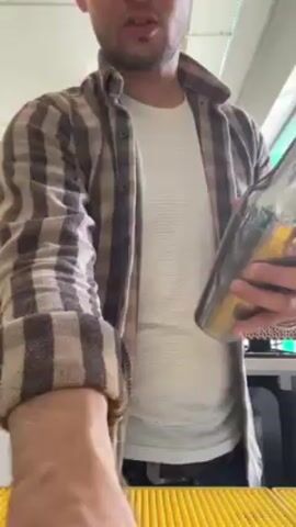 A man is making a drink. He is indoors in what looks like a bar or kitchen. He is wearing a white t-shirt, blue jeans, and a brown plaid shirt that he is not buttoning. He shakes a metal cocktail shaker with ice in it and then pours the contents of the shaker, which is a neon green liquid, into a tall glass with ice in it. The background is busy, but there is what appears to be a bar counter, kitchen appliances, and shelving. There is also music playing loudly.