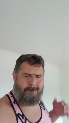 A man with a beard wearing a pink tank top with black heart and eye designs is looking at the camera and making various facial expressions and hand gestures. He is in a room with white walls and a wooden floor.