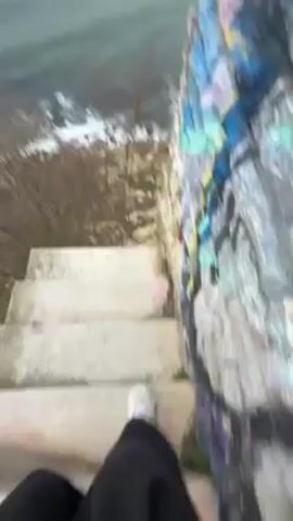 The video begins with a first-person point of view walking down a set of concrete stairs. To the right of the stairs is a concrete wall covered with colorful graffiti. At the bottom of the stairs, the camera angle moves up, displaying a coastal landscape. The sea is visible with waves crashing against the rocky shore, topped by a gray, overcast sky. There are sparse, bare trees and shrubs along the coastline, contrasting with the choppy sea. The camera continues to pan slightly, maintaining focus on the seascape. The person is wearing black pants and white shoes.