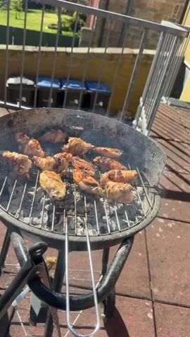A close-up shot shows chicken wings grilling on a charcoal barbecue grill outdoors. The grill is black, round, and stands on a wheeled frame on a brick patio. The chicken wings are arranged on the grill grate and appear partially cooked, some showing char marks. Smoke rises from the grill. In the background, a railing surrounds a green area, and a yellow wall and blue storage bins are visible. The camera focuses on the food and grill, showing the grilling process.