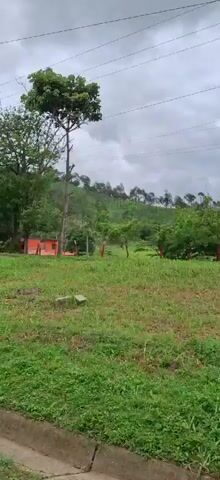 The video pans across a rural landscape on a cloudy day. In the foreground, there is green grass and a rough wooden post. In the middle ground, a small red building with an orange fence is visible, surrounded by trees. Further back, a white building with barred windows is seen. A blue motorcycle is parked near a gate with a fence. Lush green trees and hills form the backdrop. The sky is overcast.