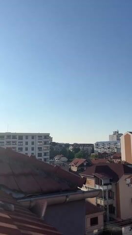 The video is a vertical shot of a cityscape under a clear blue sky. In the foreground, rooftops of houses with red tiles are visible. Beyond these, a few taller apartment buildings and other structures can be seen. The sun appears to be shining, casting light on the buildings. The overall impression is that of a quiet urban scene during the daytime.