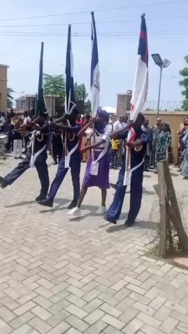 Four young people in blue and purple uniforms march in a procession holding flags. The person in the front right is holding a white flag with red and blue stripes. The person next to them on the left holds a white flag with a blue emblem. The next person holds a dark blue flag. The person on the far left holds a dark green flag. A band with brass instruments is visible in the background to the left. Other people are watching the procession from the right. The ground is paved with interlocking bricks. The sky is bright and clear.