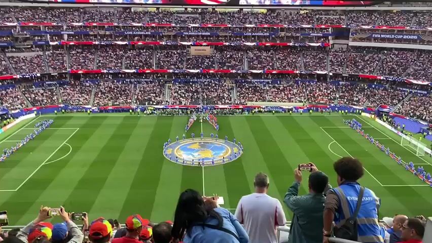 The video shows a wide shot of a packed soccer stadium during what appears to be an opening ceremony. In the center of the field, a circular stage decorated with a globe and flags is present. A line of individuals, possibly dancers or performers, are arranged around the stage. Two opposing lines of people, likely soccer players, stand on either side of the field, facing each other. The stands are filled with a large crowd of spectators, many of whom are holding up phones to record the event. The stadium seating is tiered and adorned with red, white, and blue banners. In the foreground, a few spectators are visible, including one person wearing a blue vest with "EVENT STAFF" printed on it. The overall atmosphere seems to be one of excitement and anticipation.