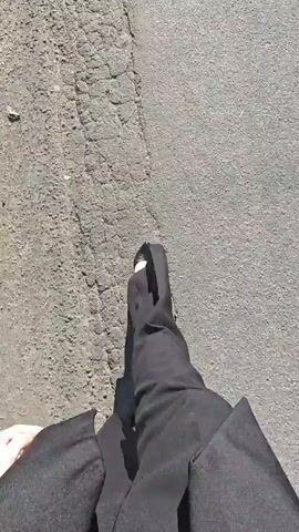 This is a first-person perspective video showing a person walking on an asphalt surface. The person is wearing black pants and black pointed-toe shoes. A dark coat or jacket is visible, draped over their legs. The ground appears to be a road or sidewalk, with some cracks and tire marks visible. The lighting suggests it is daytime.