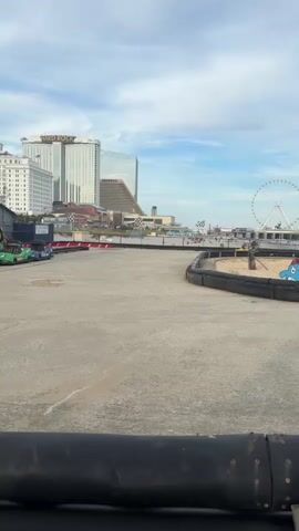 A go-kart track is shown with buildings and a Ferris wheel in the background. A child is driving a yellow go-kart around the track. Another go-kart is in the distance. The sky is partly cloudy.
