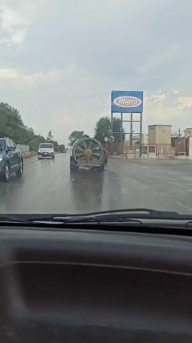 The video shows a view from inside a vehicle on a wet road. A large fan is being transported on a trailer, and several tractors are visible on the side of the road. A sign for "Migro" is in the background. The sky is overcast, and it appears to be raining.