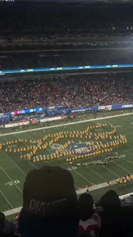 Here's a detailed description of the video:
The video shows a large crowd in a football stadium. In the center of the field, a marching band, dressed in yellow and green uniforms, is performing. They are arranged in a formation that spells out "USC". People are watching the performance from the stands. The person filming is in the stands and wearing a Giants hat. The stadium appears full. There are multiple levels of seating visible.