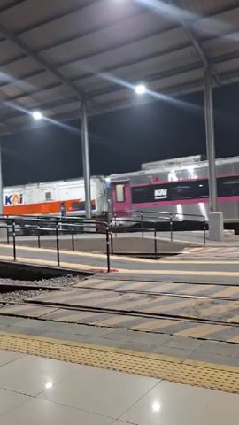 The video shows a train station at night. A train with pink and silver cars is parked at the platform. A person in a blue shirt is walking near the train. The platform has yellow and black striped markings, and there are train tracks in the foreground. The station is covered by a roof supported by pillars. Bright lights illuminate the scene.