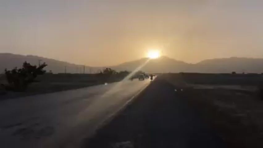 The video shows a road in a rural area at sunset. The sun is setting behind a mountain range, creating a bright glow. A motorcycle with two riders is driving down the road towards the sunset. A white car passes the motorcycle. There are some trees and power lines on the side of the road. The road is paved but appears to be a bit worn.