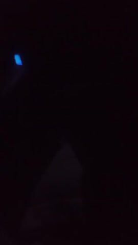 The video shows a very dark environment with a bright blue light in the upper left corner. A faint, triangular-shaped light or object is visible in the lower center of the frame. The lighting conditions suggest nighttime or an indoor setting with minimal illumination. No people or specific actions are discernible due to the darkness.
