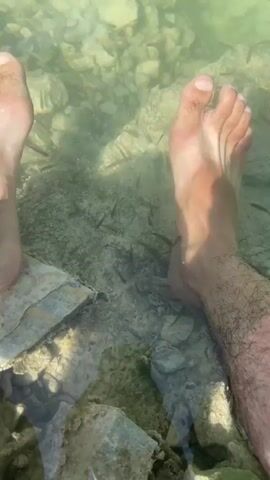 The video shows a close-up view of two bare feet submerged in clear, shallow water. Numerous small, dark fish are visible swimming around the feet and in the rocky, sandy bottom of the body of water. Sunlight filters through the water, illuminating the scene and creating dappled light patterns on the feet and the underwater environment. The focus is on the feet and the fish, with the surrounding environment appearing to be a natural outdoor setting.