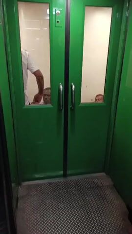 The video shows the interior of what appears to be an elevator or a similar enclosed space with green walls. The floor is covered with metal diamond plate. In the background, there are closed double doors with windows. Two people are visible through the windows of the closed doors, looking out. One person is in the foreground of the video, also appearing to be in the elevator, looking towards the closed doors. Another person is seen partially in the frame on the left side, wearing a white shirt and looking towards the doors. The lighting is dim.