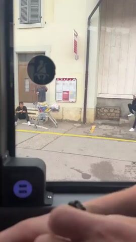 The video captures a street scene viewed from inside a vehicle, likely a bus, looking out through the window. In the foreground, a portion of the vehicle's interior is visible, including a USB charging port. Outside, two individuals are present. One person, dressed in dark clothing and white shoes, is seated on the steps of a building. Another person, wearing a hat and a dark t-shirt, stands near a shopping cart filled with items. Behind them, a light-colored building with closed shutters and a doorway is visible. A gray car is parked, partially obstructing the view. The environment appears to be a public space, possibly a street or a plaza, with yellow and blue markings on the ground indicating parking or pedestrian areas. A circular sign with a seatbelt symbol is affixed to the building. The camera perspective shifts as the vehicle moves, changing the view of the scene.